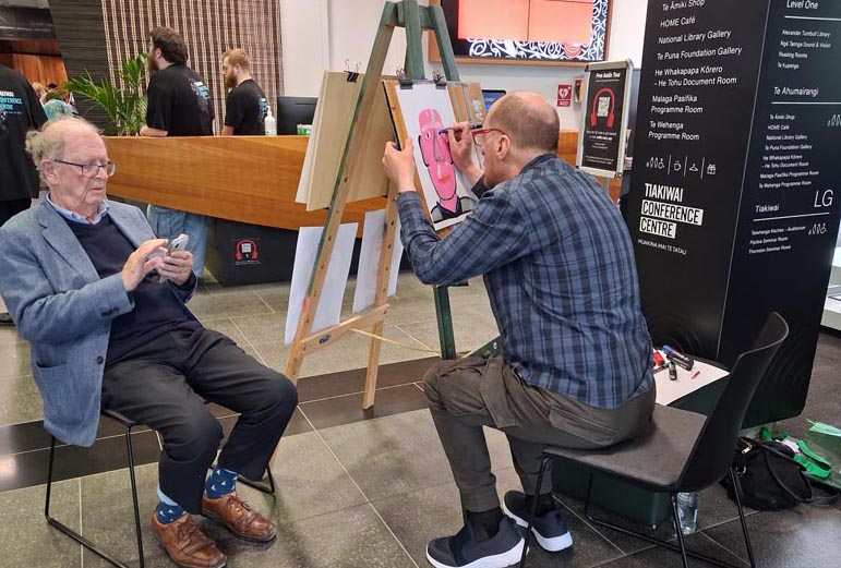 Daniel Phillips at his easel painting a portrait of a man, seated opposite